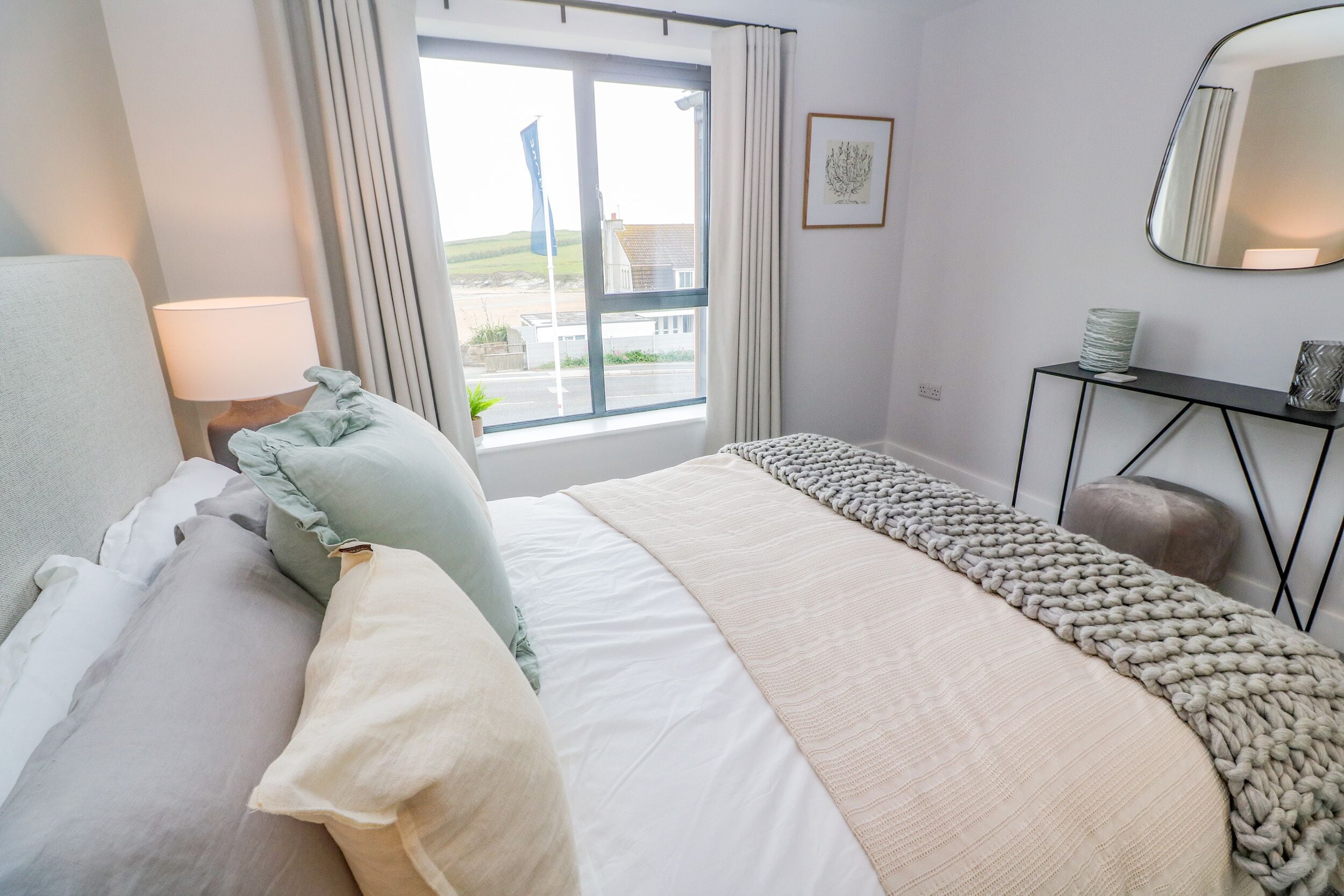 A neatly arranged bedroom with a bed, a bedside table, a mirror, and a view of the outside.