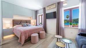 A modern bedroom with a large bed, bedside tables, a TV, and a window with a view of greenery. - Sunshine Place (Konavle)