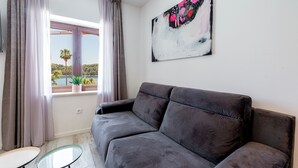 A modern living room with a grey sofa, a glass coffee table, and a large window with curtains. A painting is on the wall. - Sunshine Place (Konavle)