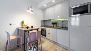 A modern kitchen with white cabinets, a stainless steel refrigerator, and a dining area with two pink chairs and a table set with fruits and drinks. - Sunshine Place (Konavle)