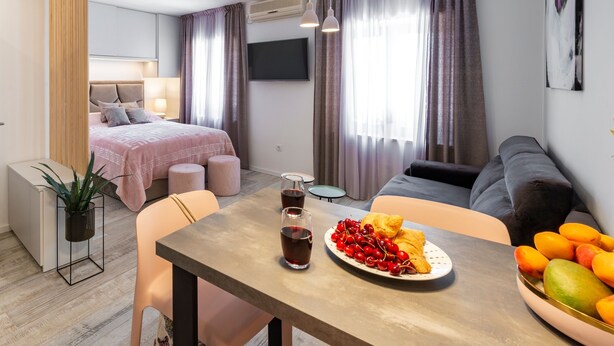 A modern hotel room with a bed, a sofa, a dining table with fruit and drinks, and a TV on the wall. - Sunshine Place (Konavle)