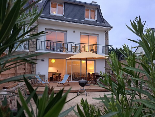 Una casa de dos pisos con balcón, un patio con mobiliario exterior y una parrilla.