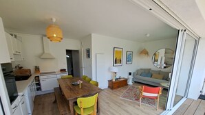 A modern kitchen with white cabinets, a wooden dining table, and a yellow chair. There is a sofa and a coffee table in the living area, and a large window with a view of the outside.