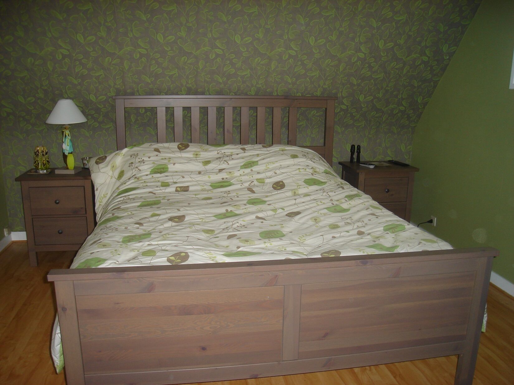 A bedroom with a wooden bed, green patterned wallpaper, a nightstand with a lamp, and a dresser.