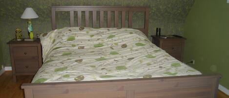 A bedroom with a wooden bed, green patterned wallpaper, a nightstand with a lamp, and a dresser.