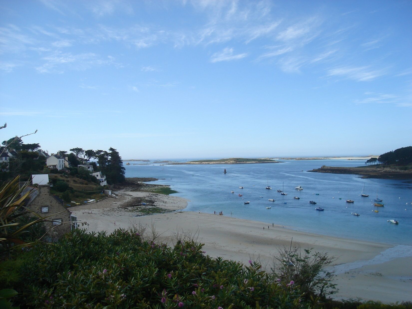 Vista costera con playa de arena, barcos en el agua y casas a la orilla.