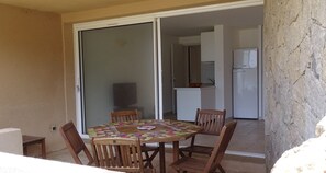 Un coin repas avec une table ronde et quatre chaises, une porte-fenêtre coulissante donnant sur une cuisine et un réfrigérateur blanc.
