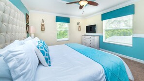 A bedroom with a tufted headboard, a bed with white and blue bedding, a dresser, a television, and a ceiling fan.