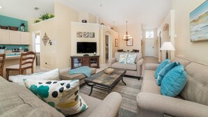 A living room with a sofa, coffee table, TV, and kitchen area with cabinets and a dining table. - Last Minute Disney! Private Pool, Hot Tub & Themed Bedrooms. Book Now! #21748 (Orlando)