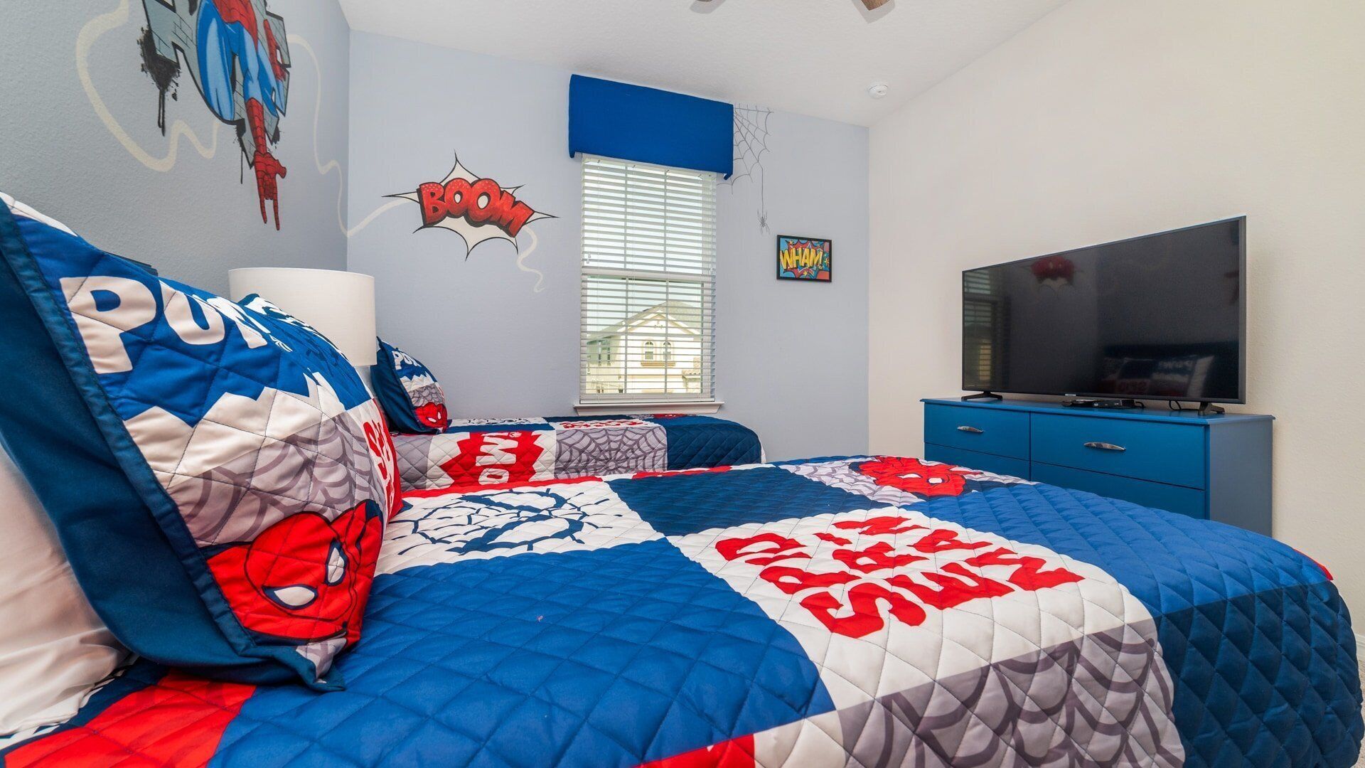 Et soveværelse med en seng dækket af et quilt med Spider-Man motiver, en blå kommode, et fjernsyn og et vindue med persienner.