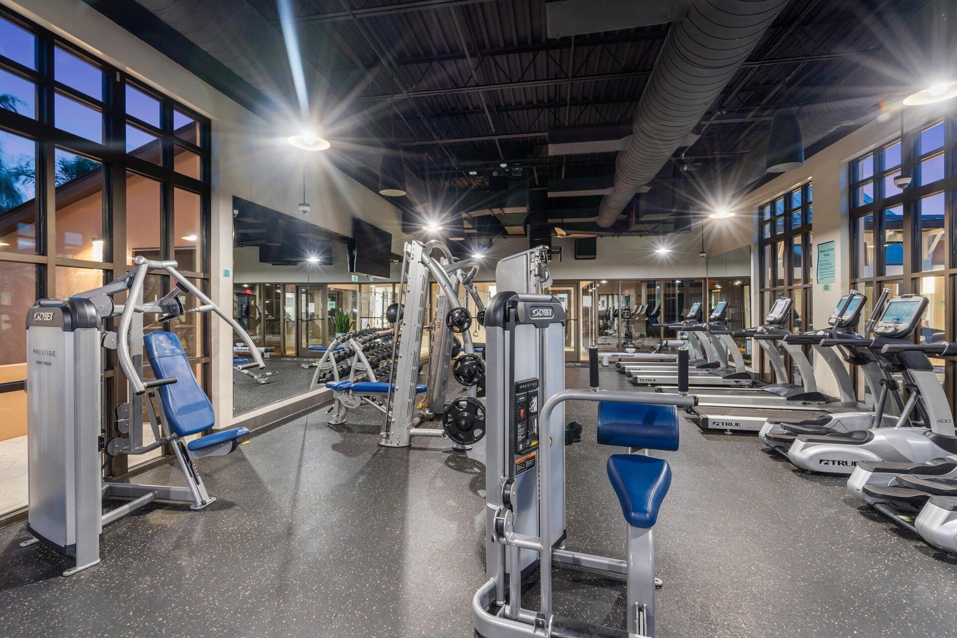 Un gimnasio con diversos equipos de ejercicio, amplias ventanas y un techo alto.