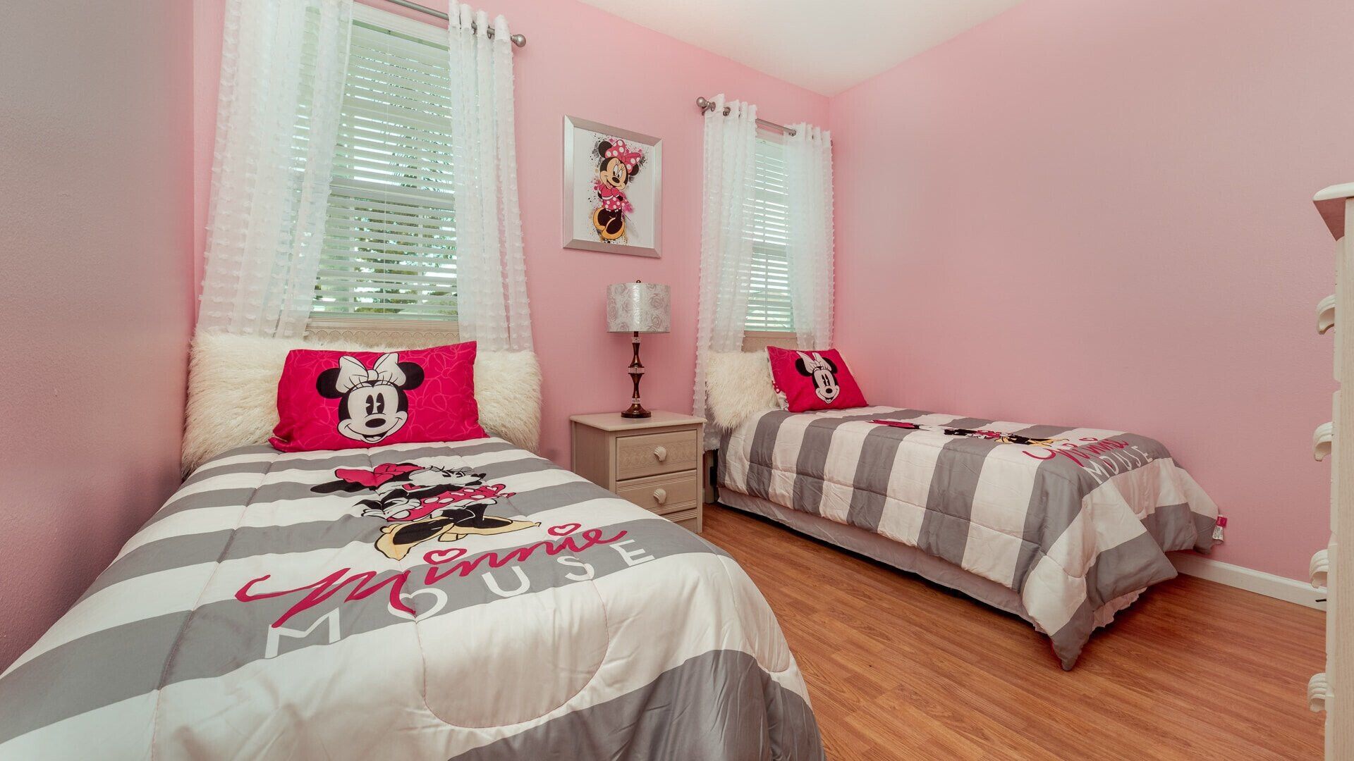 A bedroom with two beds, each with a Minnie Mouse themed bedspread, a nightstand with a lamp, and a picture on the wall.