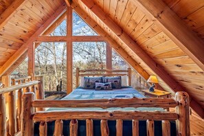 Une chambre à coucher aménagée dans les combles, avec un grand balcon, un lit recouvert d’une couette aux motifs forestiers et une rambarde en bois.