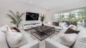 Una sala de estar moderna con televisor de pantalla plana, sofás blancos, una mesa de centro y ventanas amplias.