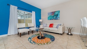 Una sala de estar con una pared azul decorativa, un sofá blanco, una mesa de centro de vidrio y un tapete colorido.