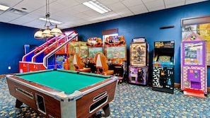 Una mesa de billar en una sala con juegos de arcade, una escalera y una alfombra estampada.