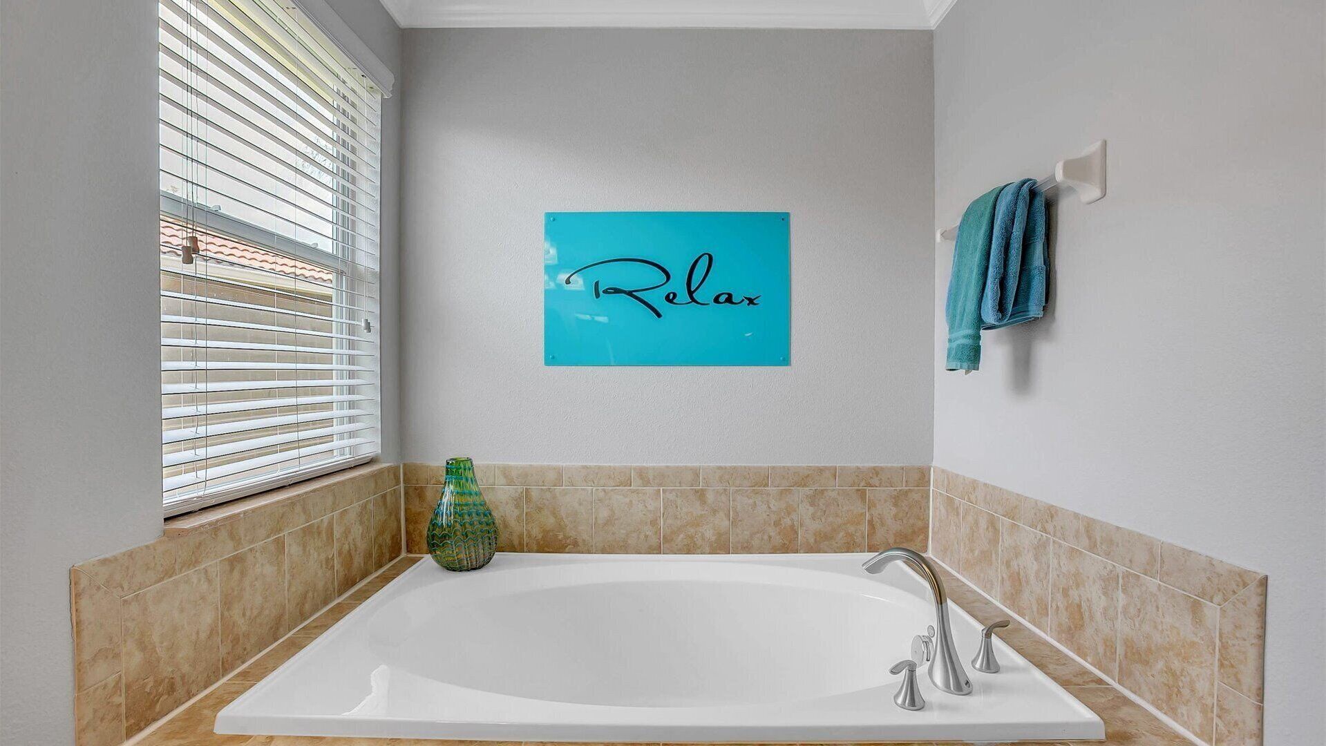 A bathroom with a bathtub, a towel rack with towels, a window with blinds, and a 'Relax' sign on the wall.