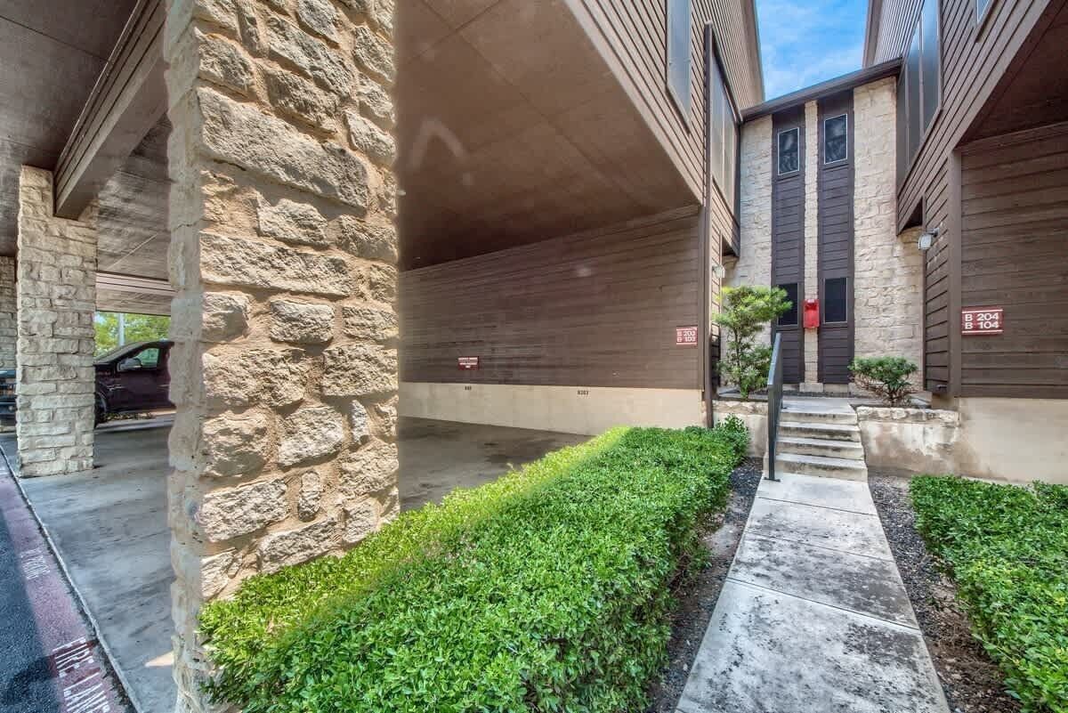 A covered parking area with stone pillars, a concrete floor, and a small garden with a pathway leading to a building entrance.