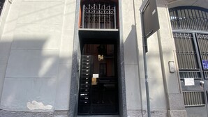 Ein Gebäudeeingang mit einem schwarzen Metallgitter, einer Treppe, die zum Eingang führt, und einem kleinen Schild darüber.