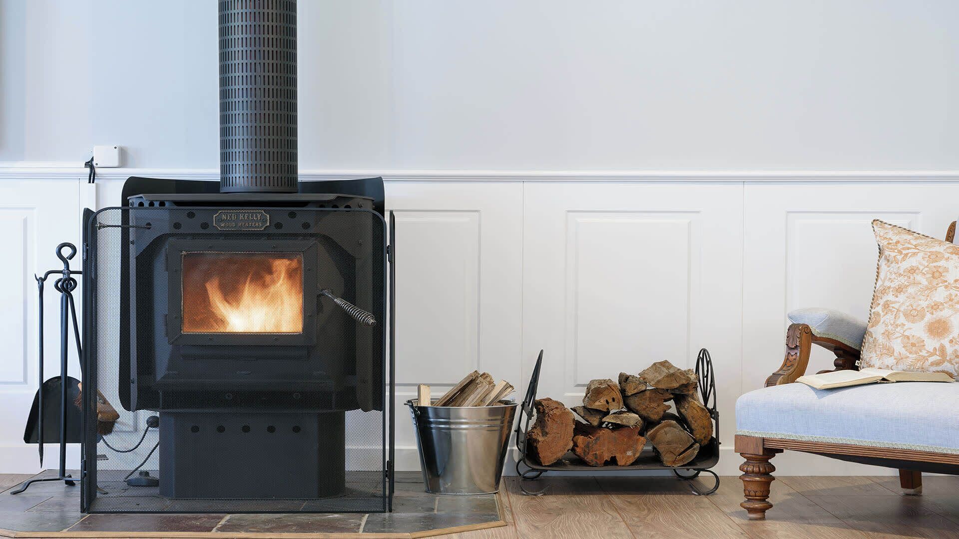 A wood-burning stove with a metal grate, a metal bucket of firewood, and a chair with a cushion.