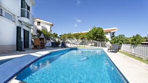 Une piscine rectangulaire à l’eau d’un bleu limpide, entourée d’une clôture blanche et jouxtant un bâtiment doté de balcons et d’un espace de détente extérieur.