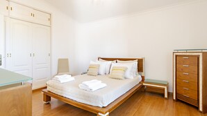 Une chambre à coucher bien rangée, avec un lit en bois, du linge de lit blanc, un banc, une armoire et une table en verre.