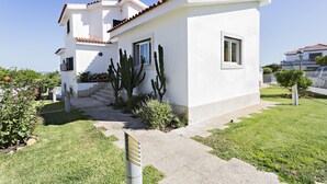 Un bâtiment blanc avec un toit recouvert de tuiles, un petit jardin agrémenté de cactus et de fleurs, et un ciel d’un bleu limpide.
