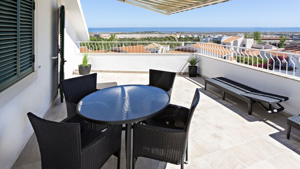 Un balcon avec une table ronde et des chaises, un banc, et une vue sur la mer et les bâtiments.
