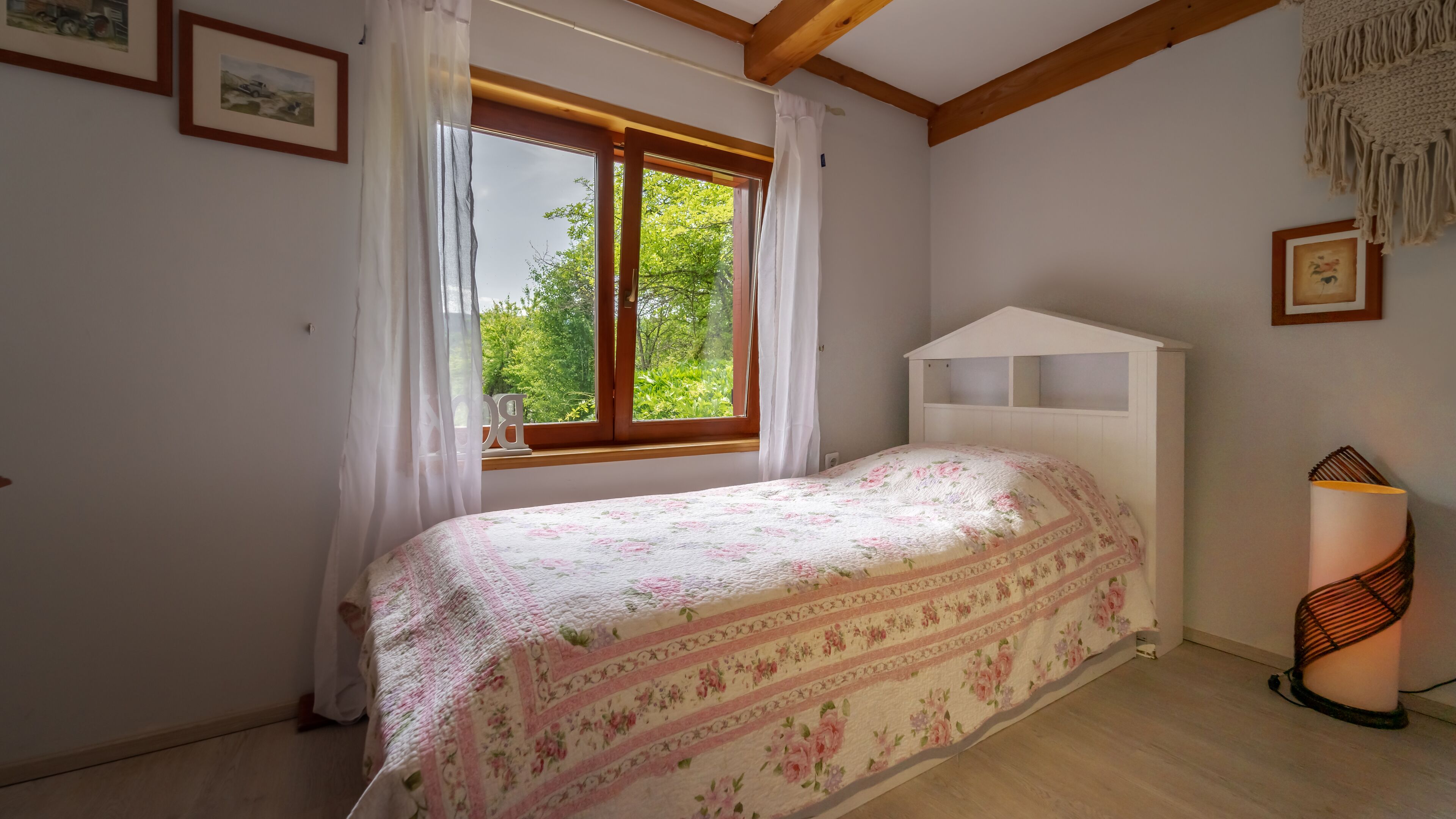 A bedroom with a bed, a window with curtains, a bedside lamp, and framed pictures on the wall.