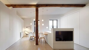 A modern living room with a built-in TV, wooden flooring, and a dining area with white chairs and a wooden table. - Luna by FeelFree Rentals (San Sebastian)