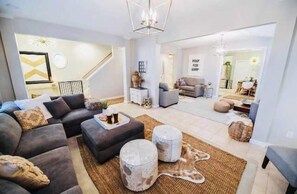 A modern living room with a sectional sofa, ottoman, and round coffee table. There is a staircase leading to another level, and a dining area visible in the background.