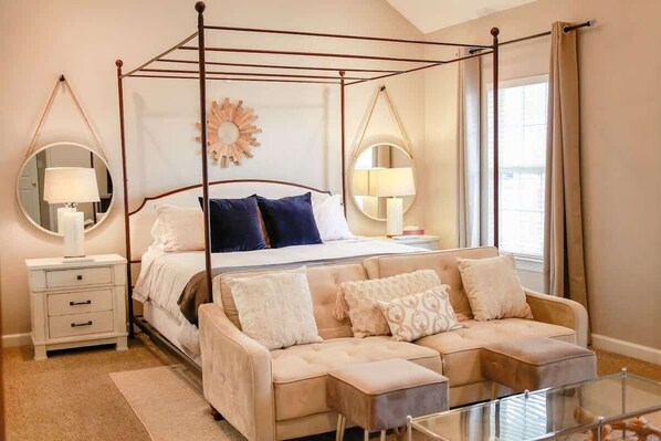 A four-poster bed with a mirror, bedside tables, a sofa, and a glass coffee table in a well-lit room with large windows.