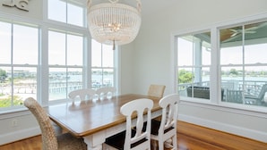 Un espace repas avec une table en bois et des chaises blanches, de grandes fenêtres donnant sur le front de mer et un lustre.