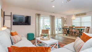 A modern living room with a sofa, coffee table, and a TV mounted on the wall. There is a dining area with a table and chairs, and a sign that reads "BEACH HOUSE". - Newly renovated home across from the beach! (Carolina Beach)