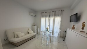 Een moderne woonkamer met een bank, een glazen eettafel, transparante stoelen, een kast en een aan de muur bevestigde televisie.