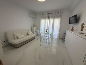 Ein modernes Wohnzimmer mit einer Couch, einem Glastisch, transparenten Stühlen, einem Schrank und einem an der Wand befestigten Fernseher.