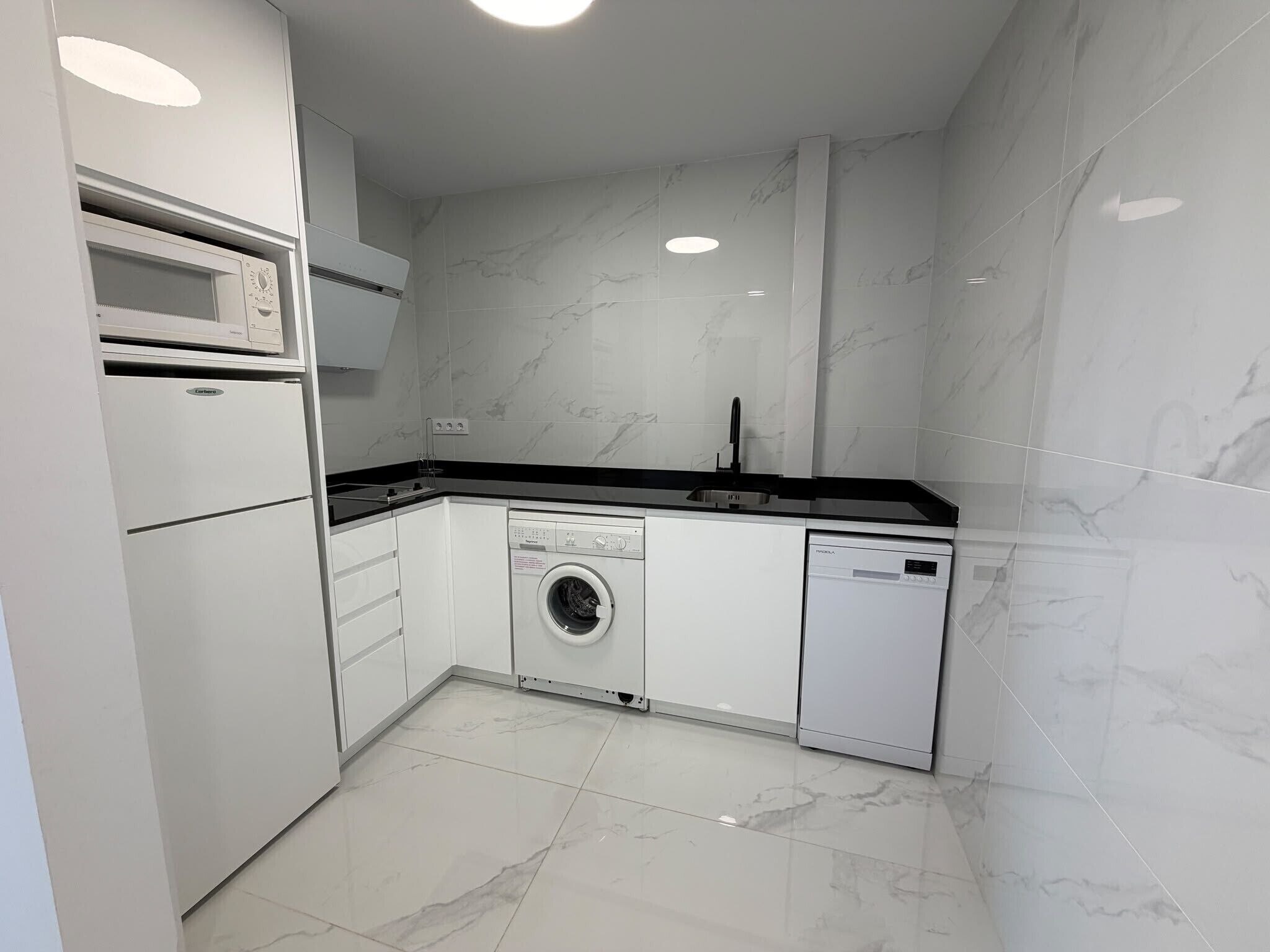 Cocina compacta con lavadora, lavavajillas, microondas y un lavabo con encimera negra.