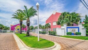 A well-maintained residential area with a paved driveway, palm trees, and a red house with a blue gate.