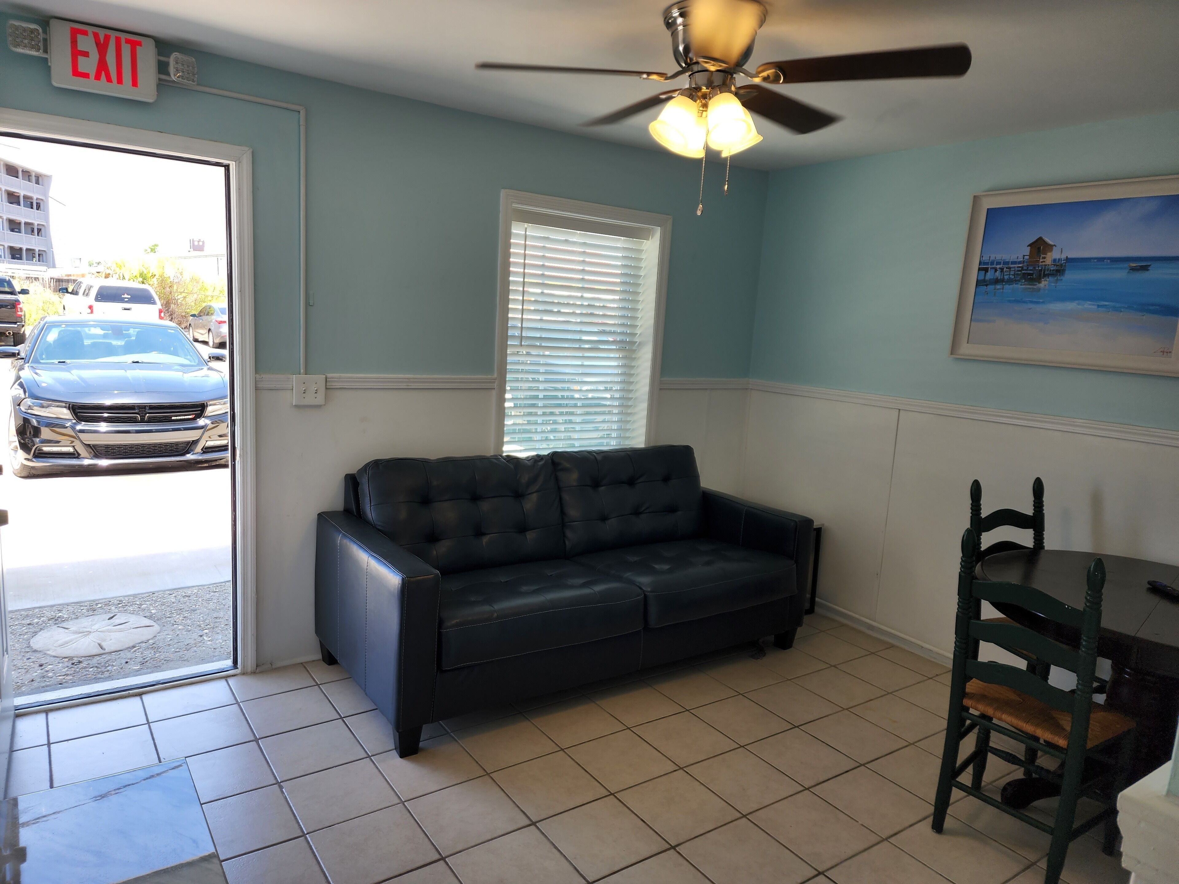 A room with a black leather sofa, a dining table with a chair, a ceiling fan, a window with blinds, and an exit sign.