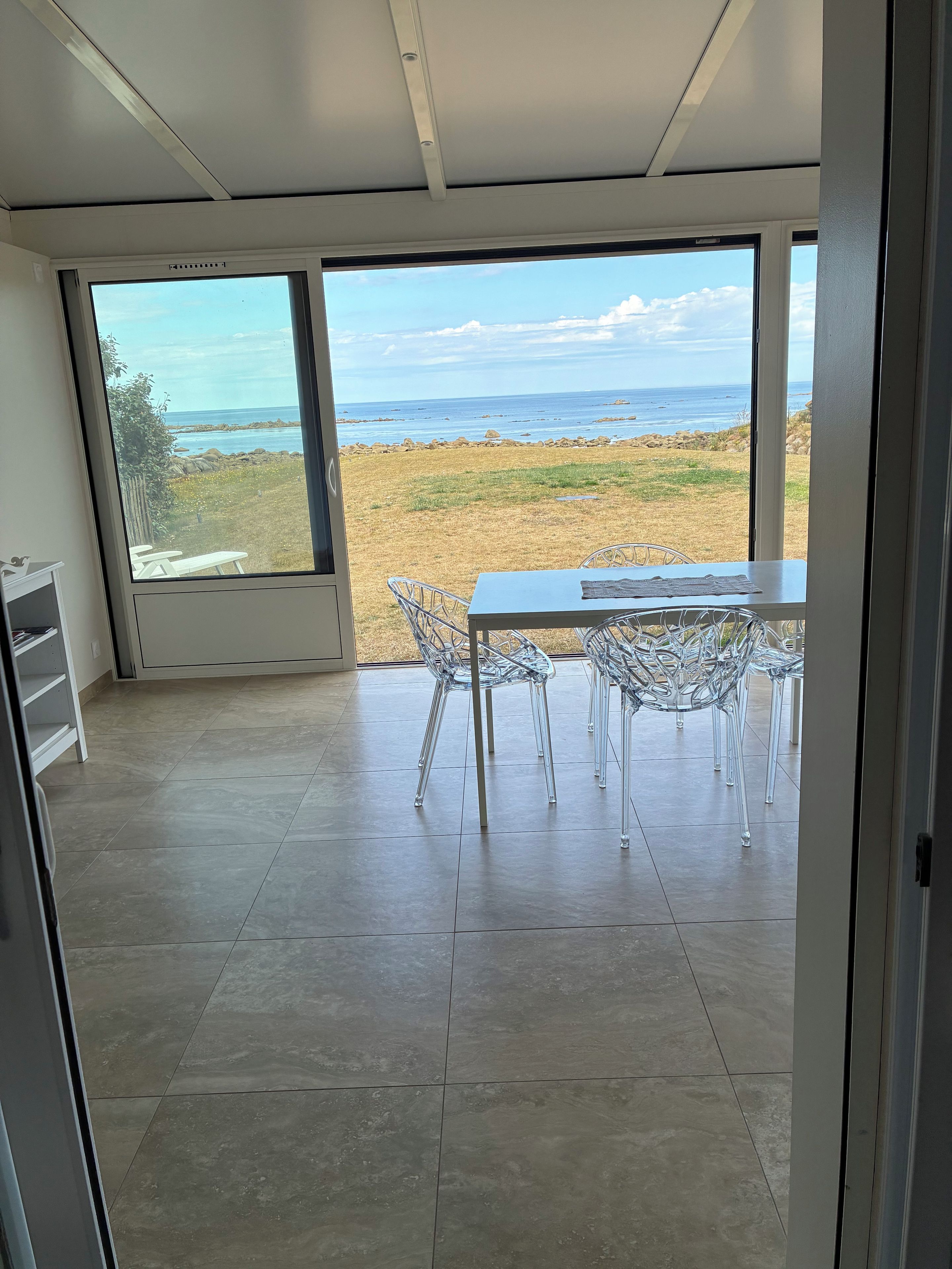 A room with a large window overlooking a beach, a table with chairs, and a tiled floor.