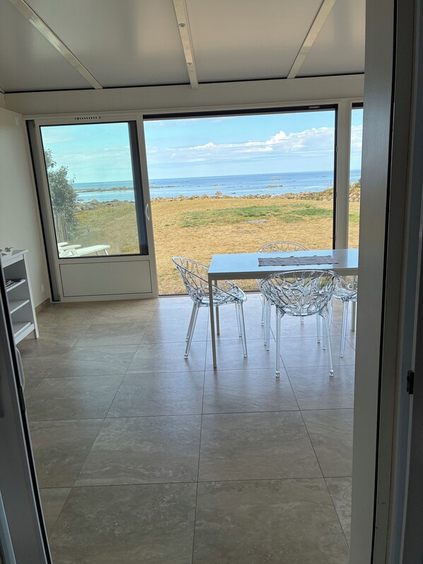 A room with a large window overlooking a beach, a table with chairs, and a tiled floor.