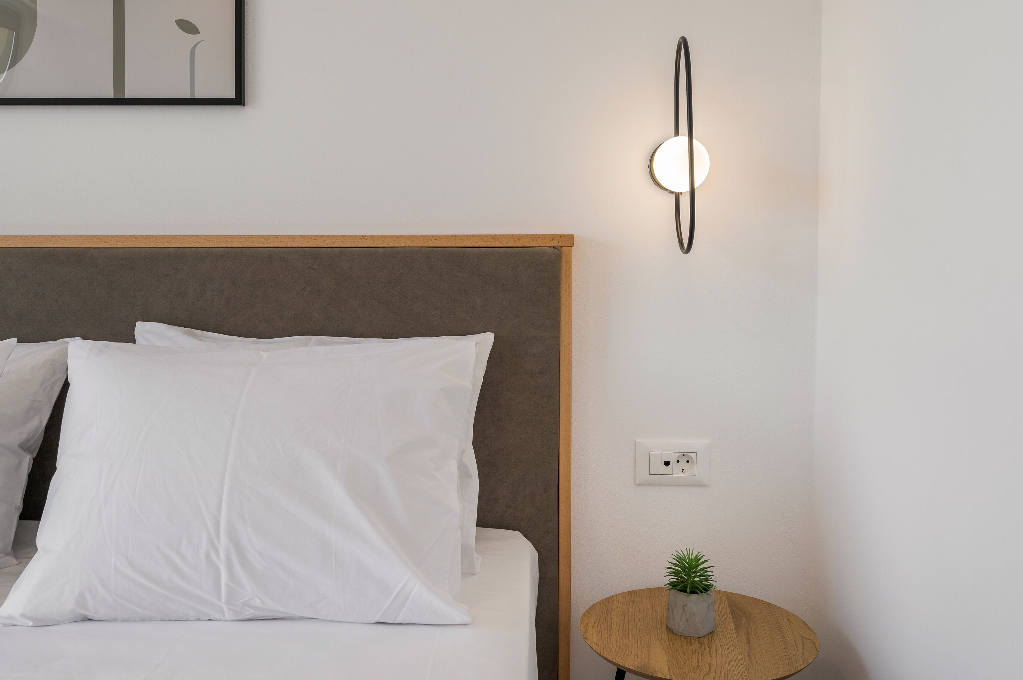 A neatly made bed with white bedding and a small round table with a plant beside it.