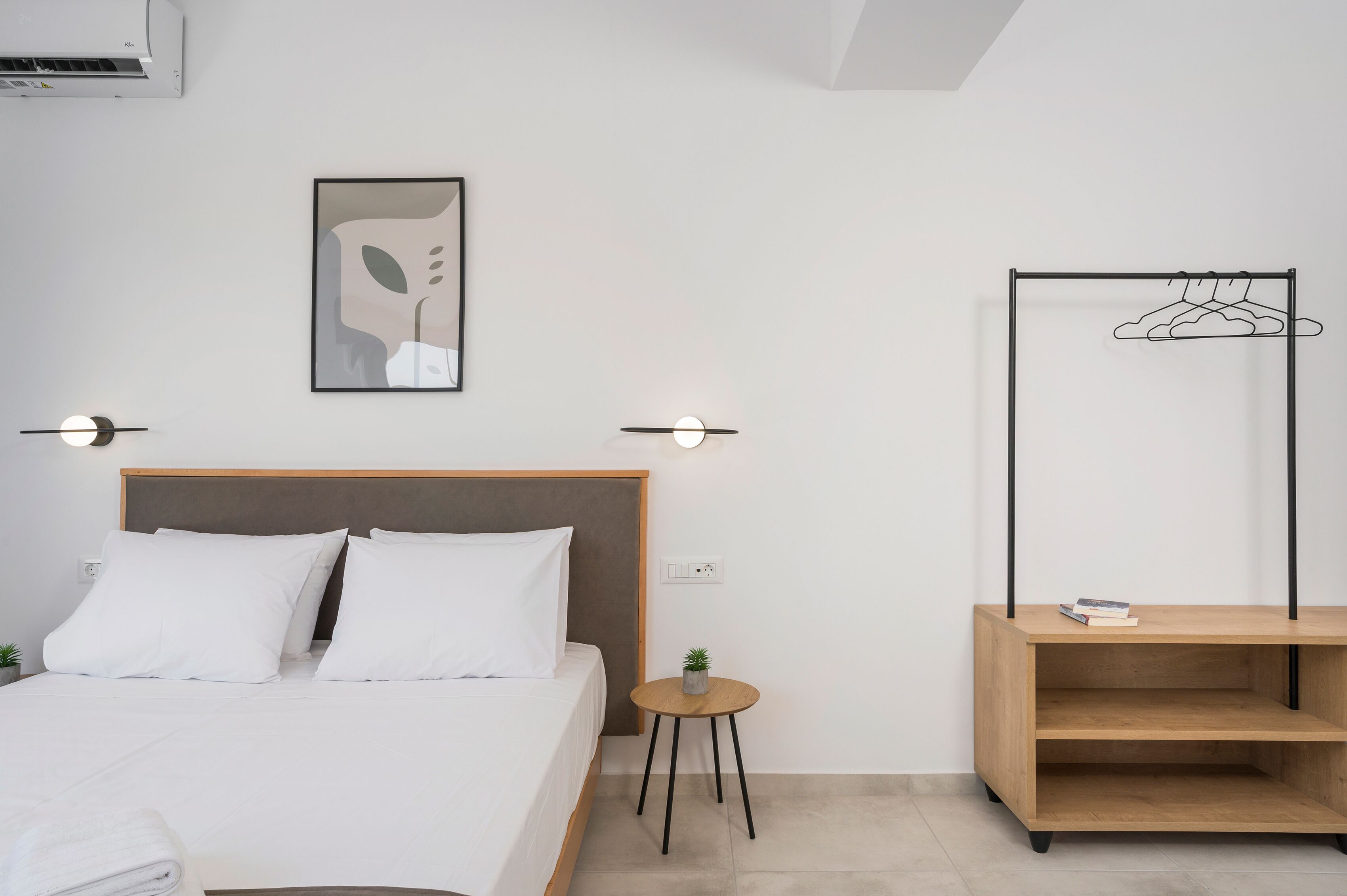 A modern hotel room with a bed, bedside table, wall-mounted air conditioner, abstract artwork, wall-mounted light, and a wooden shelf with a hanger.