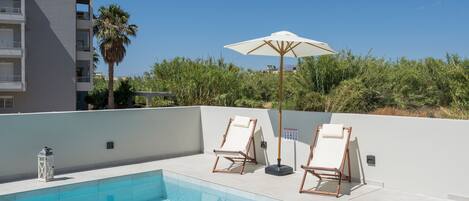 Zona de piscina en la azotea con dos sillones de relax y una sombrilla grande, rodeada de vegetación y un edificio al fondo.