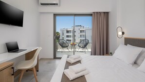 Una habitación de hotel moderna con una televisión de pantalla plana, un escritorio con una computadora portátil, una cama con ropa blanca, dos sillas, un balcón con vistas a edificios y un equipo de aire acondicionado.