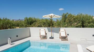Une piscine avec deux chaises longues et un parasol, entourée de verdure et sous un ciel dégagé.