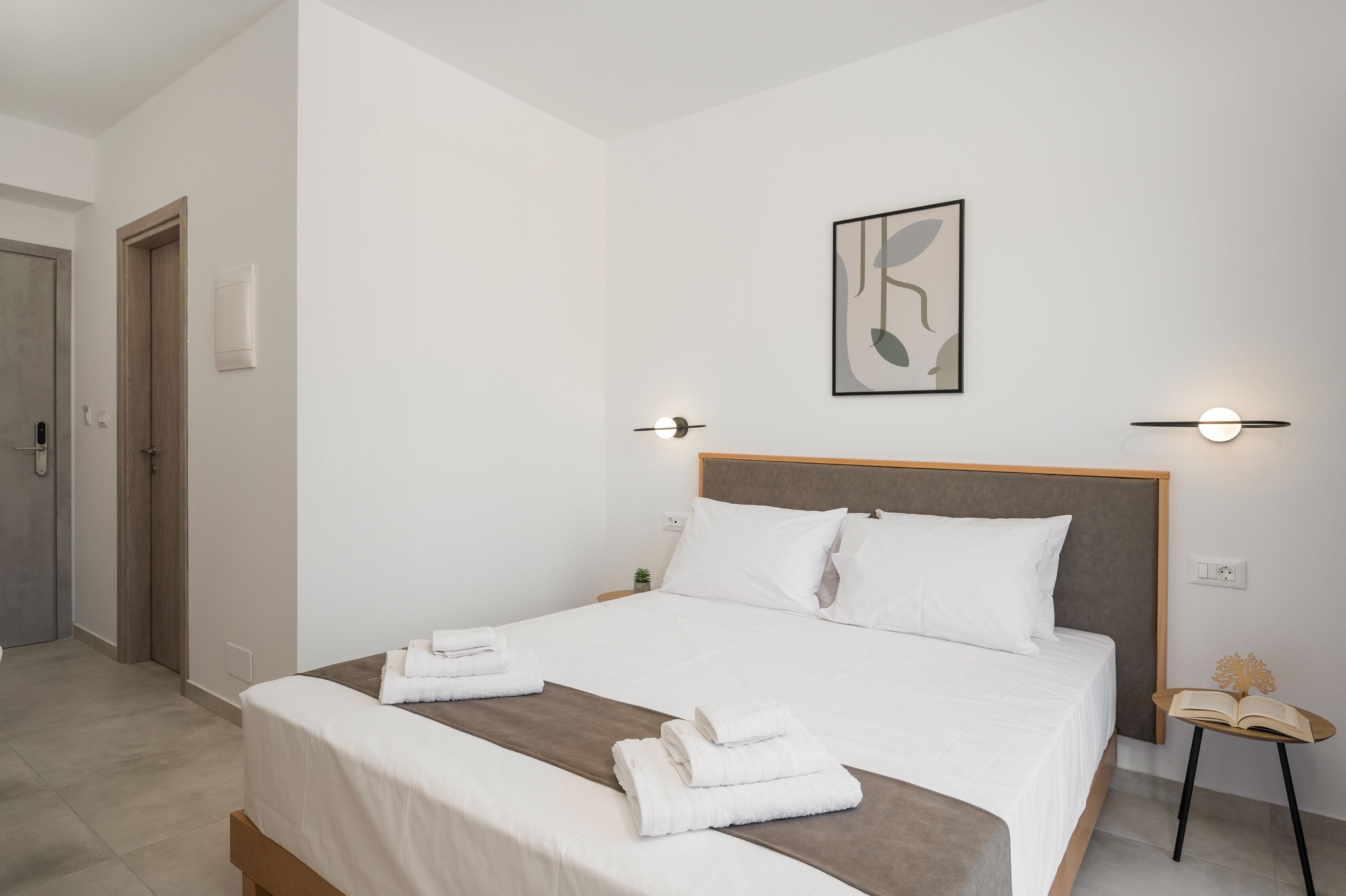Una cama bien hecha con ropa de cama blanca y un detalle marrón, una mesita de noche con un libro y una planta pequeña, y una obra de arte colgada en la pared.