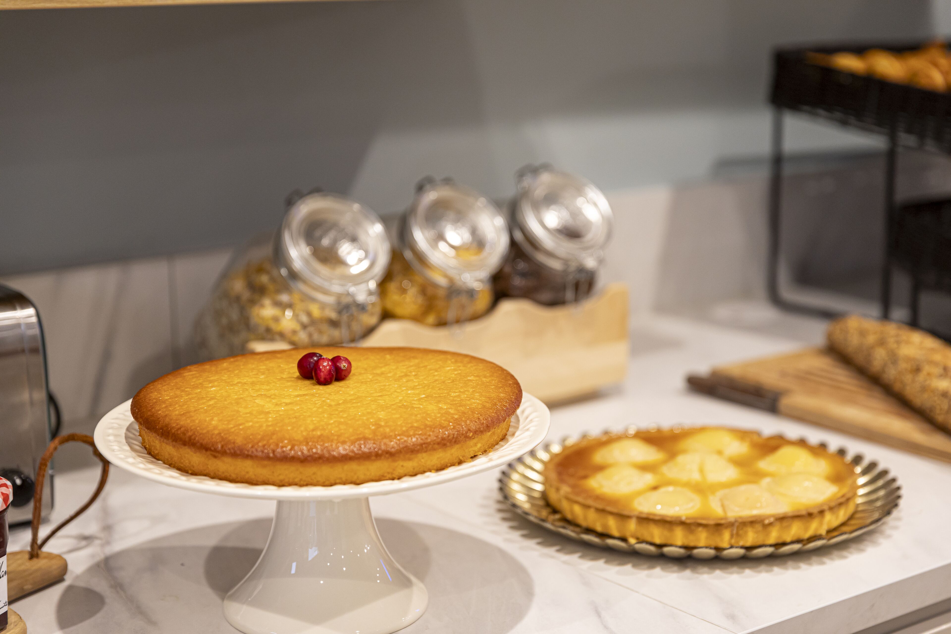 A cake on a white pedestal stand with a cherry on top, a pie on a round plate, and jars of jam in the background.