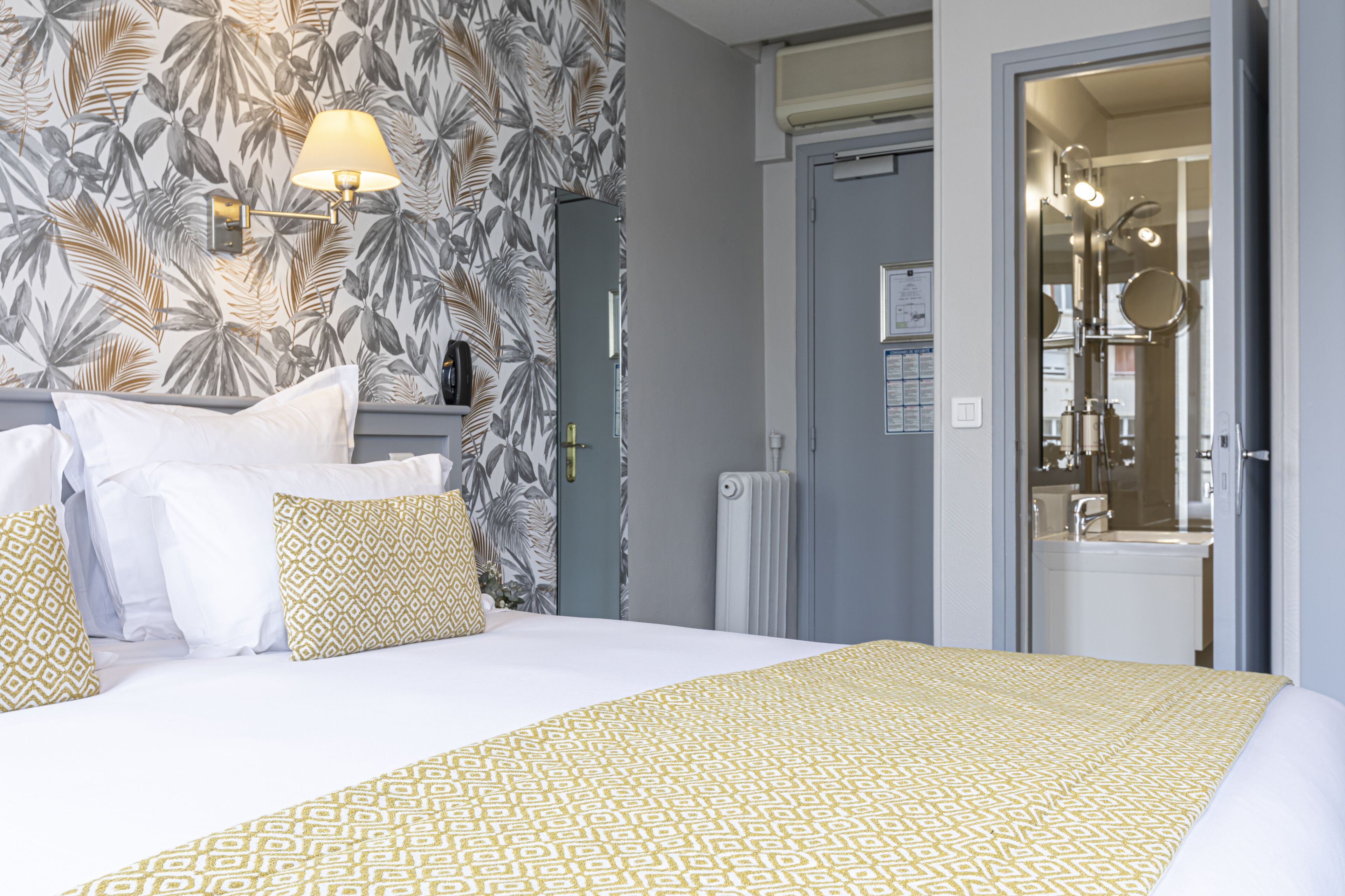 A hotel room with a bed, a nightstand, a lamp, a mirror, and a door leading to a bathroom.