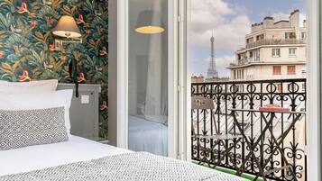 Habitación de hotel con cama, almohadas estampadas, mesita de noche, lámpara y vista de la Torre Eiffel a través de un balcón con barandilla de hierro forjado.